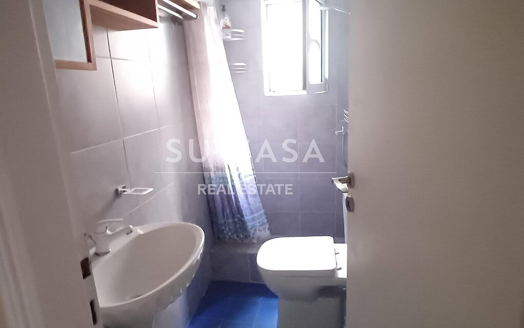 Μικρό μπάνιο με λευκά πλακάκια, νιπτήρα και παράθυρο, σε διαμέρισμα στην Ελλάδα.